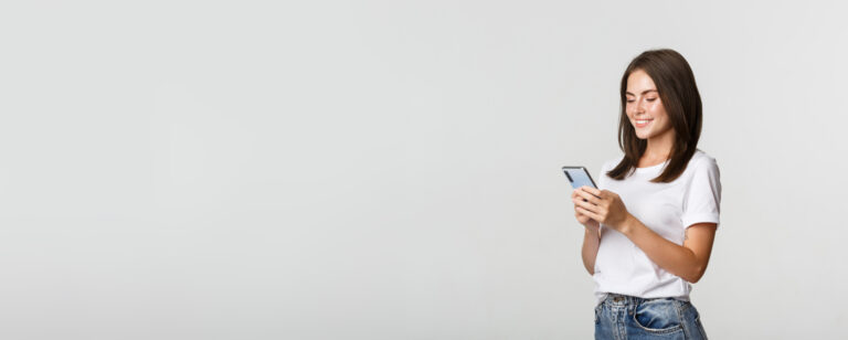 Junge Frau mit braunen Haaren lächelt und schaut auf Smartphone, trägt weißes T-Shirt, blaue Jeans.