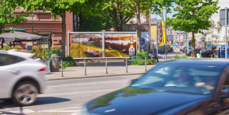Eine belebte Straße mit vorbeifahrenden Autos und einer bunten Werbetafel im Hintergrund