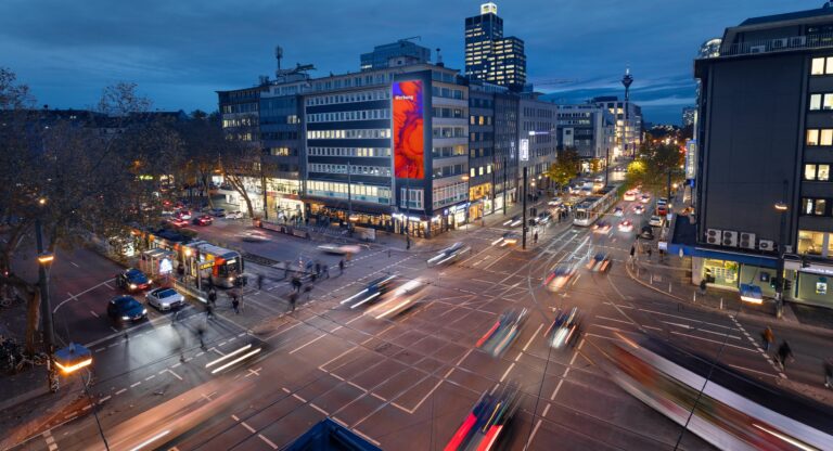 Belebte Straßenecke bei Dämmerung mit bunt beleuchteter Gebäudefassade und hellem Himmel