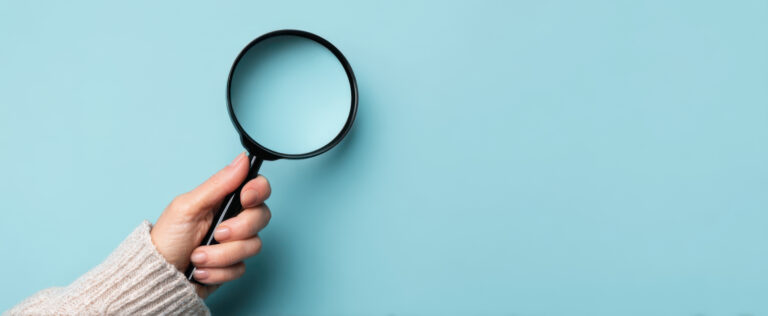 Eine Hand in hellem Pulli hält eine Lupe vor blauem Hintergrund