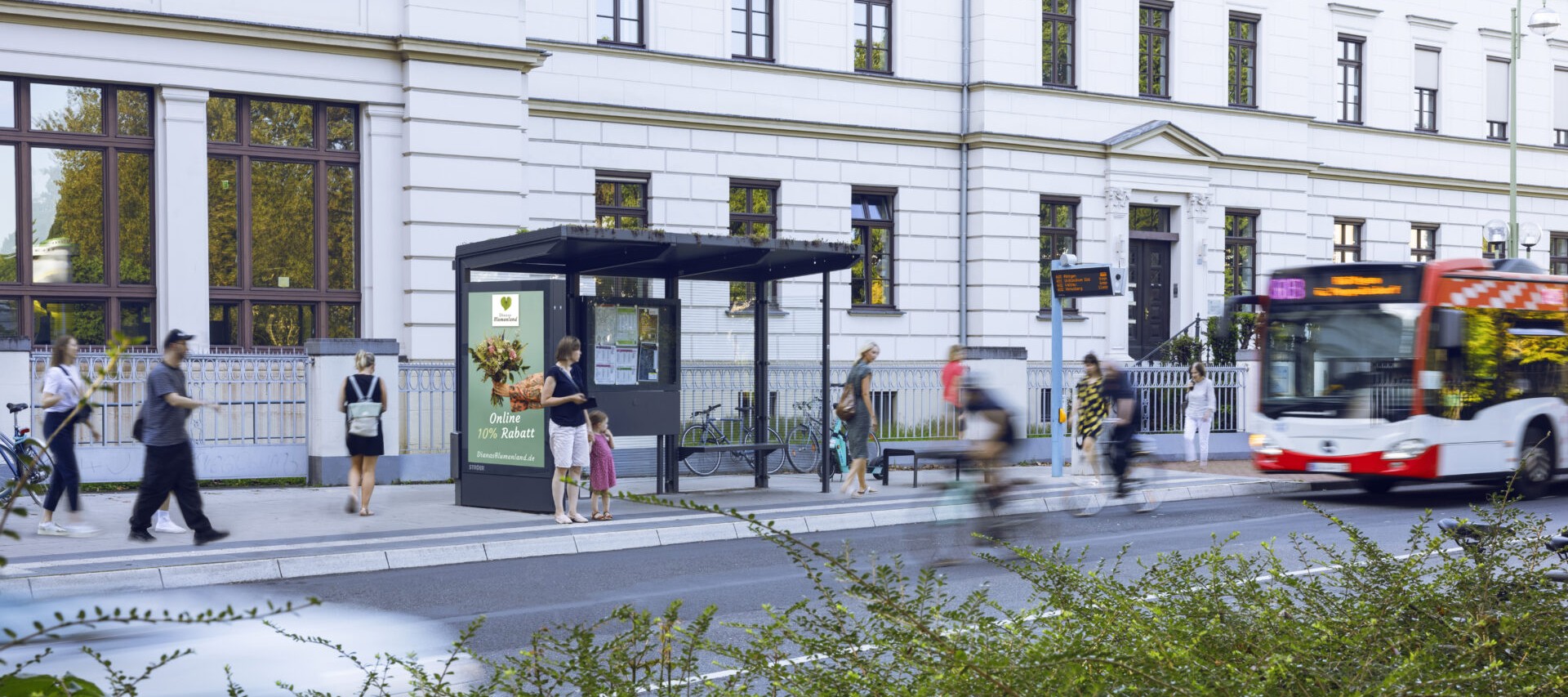 An einer belebten Bushaltestelle warten mehrere Menschen bei sonnigem Wetter
