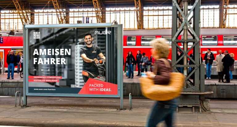 Ein junger Mann mit dunklen Haaren lacht freundlich auf einem Werbeplakat mit rotem Akzent