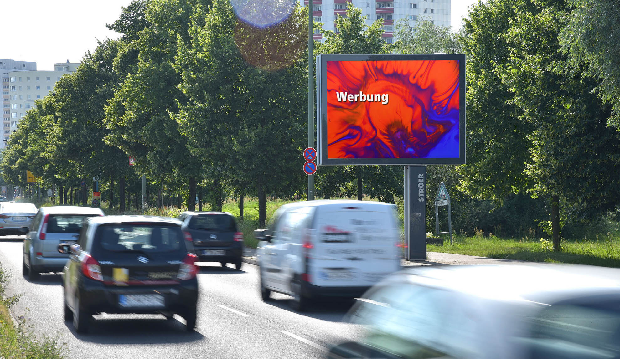 Mehrspurige Straße mit fahrenden Autos und rotem Werbeplakat vor grünen Bäumen