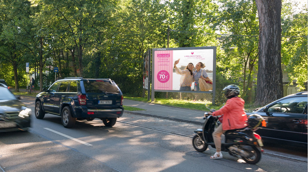 Zwei lächelnde Personen auf pinkweißem Werbeplakat am Waldstraßenrand