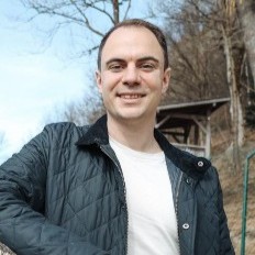 Laechelnder Mann in schwarzer Jacke und weissem Shirt im Freien