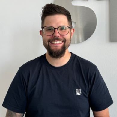 Lachender Mann mit Brille Bart dunkelblauem Shirt und Logo vor Grau